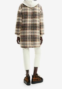 Cappotto a quadri in crema, nero e marrone con cappuccio; abbinato a pantaloni bianchi a tre quarti e scarpe nere con dettagli a motivo check arancione.