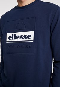 Muž v námořnickém modrém svršku se zdobeným a vytištěným logem ellesse a textem "Založeno 1959" na hrudi.