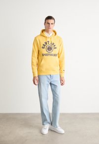 Sweatshirt à capuche jaune avec texte bleu foncé, poche avant, porté avec un jean bleu clair et des baskets blanches. Design simple et décontracté.