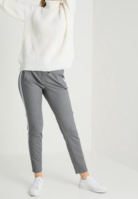 Pull à col roulé en tricot blanc, pantalon gris slim avec des rayures sur les côtés, et baskets blanches. Texture douce et design décontracté.