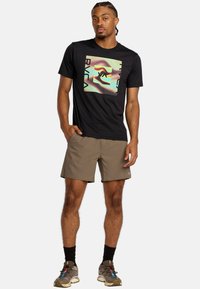 Schwarzes T-Shirt mit kurzen Ärmeln und grafischem Design, kombiniert mit beigen Shorts. Zubehör umfasst schwarze Socken und mehrfarbige Turnschuhe.