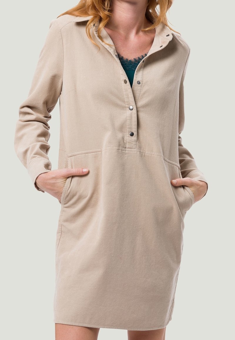 Femme portant une robe chemise beige à manches longues avec boutons-pression et poches avant, les mains posées de manière décontractée dans les poches.
