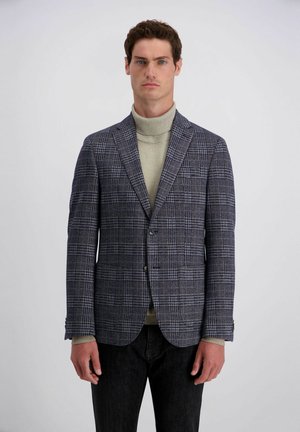 Mann trägt einen grauen karierten Blazer über einem beigen Rollkragenpullover und schwarzen Hosen, steht vor einem schlichten hellen Hintergrund.