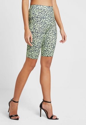 Ben iført grønne biker shorts med leopardprint og sorte højhælede sandaler med ankremod en ensfarvet baggrund.