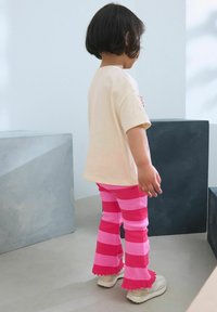 Kind met een crèmekleurig T-shirt en felroze gestreepte flare-broek met een golfrand. Schoenen zijn licht van kleur met een chunky design.