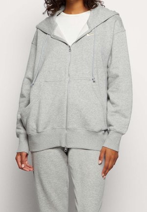 Personne portant un sweat à capuche gris clair zippé avec des cordons et un pantalon de jogging assorti, laissant apparaître partiellement un t-shirt blanc en dessous.