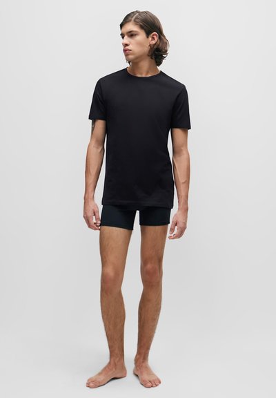 T-shirt noir à manches courtes avec un col rond, en tissu lisse. Porté avec un short noir ajusté. Le modèle se tient pieds nus contre un fond uni.