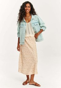 Džínová bunda v svetlomodrej farbe, biela háčkovaná blúzka, béžová maxi sukňa s kvetinovým vzorom, hnedé ploché sandále, dlhé kučeravé vlasy.