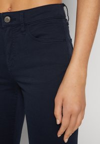 Námornícky modré džínsy z denimu s hladkou textúrou, s stredne vysokým pásom a kovovým zapínaním. Viditeľné stehy na vreckách a úzky strih.
