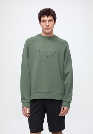 Homme aux cheveux bouclés portant un sweat-shirt vert Ted Baker Sport et un short noir, debout devant un fond clair uni.