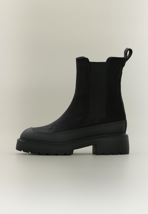 Bota Chelsea negra con paneles laterales elásticos, lengüeta en el talón, suela gruesa con taco y tacón bajo en bloque sobre fondo neutro.