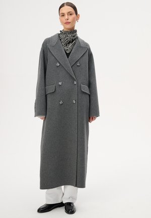 Kobieta w długim, szarym, dwurzędowym płaszczu, czarno-białym szaliku w wzory, białych spodniach i czarnych butach, stojąca na jednolitym tle.