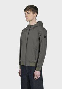 Nohavična jakna maslinaste boje s kapuljačom, prednjim patentnim zatvaračem, elastičnim manžetama i donjim rubom; ima bočni džep s patentnim zatvaračem i logotip na rukavu.