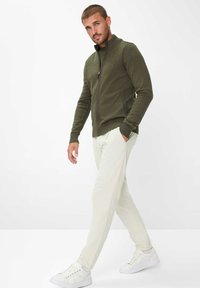 Olivgrüner Strickpullover mit Reißverschluss, gerippten Bündchen, kombiniert mit hellbeigen Cordhosen und weißen Turnschuhen. Schlichtes Design, ohne Muster.