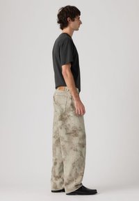 Φωτεινά καφέ τζιν με εφέ tie-dye και άνετη γραμμή, σε συνδυασμό με κοντομάνικη μπλούζα σκούρου γκρι. Μαύρα παπούτσια ολοκληρώνουν την εμφάνιση.