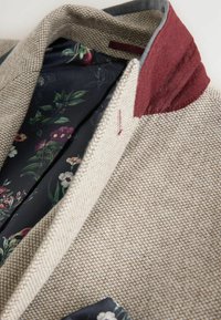 Blazer beige texturizado con un cuello burdeos. El interior presenta un patrón floral sobre un tejido oscuro. Costuras detalladas visibles en la solapa.