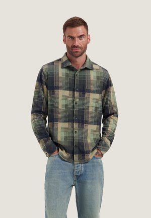 Uomo con capelli castani corti che indossa una camicia a quadri verde e blu navy abbottonata e jeans azzurri, mani in tasca, volto in avanti.