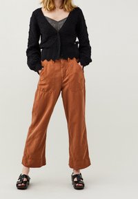 Czarny dziergany kardigan z fakturowanym wzorem, zestawiony z rudymi lnianymi spodniami o dużych kieszeniach i luźnym kroju. Czarne sandały.