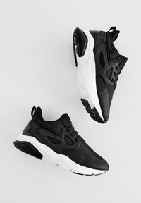 Baskets de sport noires avec une tige texturée, semelle intermédiaire blanche et semelle extérieure en caoutchouc noire. Présente des superpositions courbées et une languette au niveau du talon.
