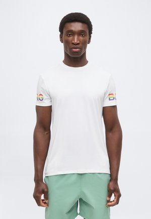 Mężczyzna ubrany w białą koszulkę z tęczowym napisem "BLG" na rękawach i jasnozielone szorty, stojący na jednolitym jasnym tle.