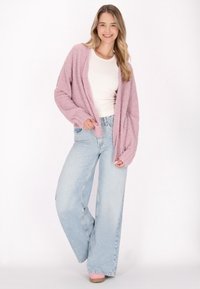 Ljusrosa fuzzy cardigan över en vit topp, kombinerad med ljusblå vida jeans och ljusrosa klackar. Mjuk textur och avslappnad passform.