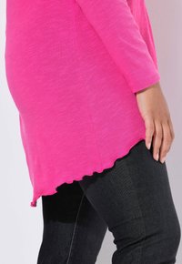 Maglietta a maniche lunghe di un vivace colore rosa con orlo smerlato, realizzata in un tessuto testurizzato, abbinata a jeans in denim neri. Primo piano sui dettagli del design.
