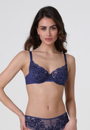 Donna che indossa reggiseno e mutandine in pizzo blu navy coordinati con motivi floreali, in piedi davanti a uno sfondo chiaro e uniforme.