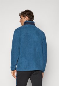 Blå fleecejakke med høy krage, med en teksturert overflate og synlige sømmer. Designet inkluderer lange ermer og en avslappet passform.