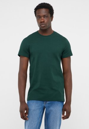 Junger Mann steht still, trägt ein dunkelgrünes Kurzarmhemd und hellblaue Jeans vor einem einfarbigen hellen Hintergrund.