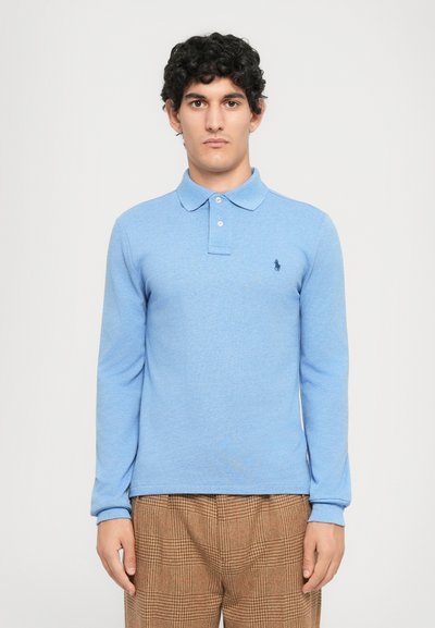 Un jeune homme portant une chemise polo à manches longues bleu clair et un pantalon à carreaux marron, se tenant devant un fond uni clair.