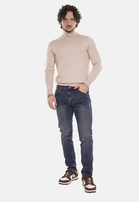 Pullover beige a collo alto, aderente, abbinato a jeans slim blu scuro. Sneakers marroni con dettagli bianchi. Outfit casual e con texture.