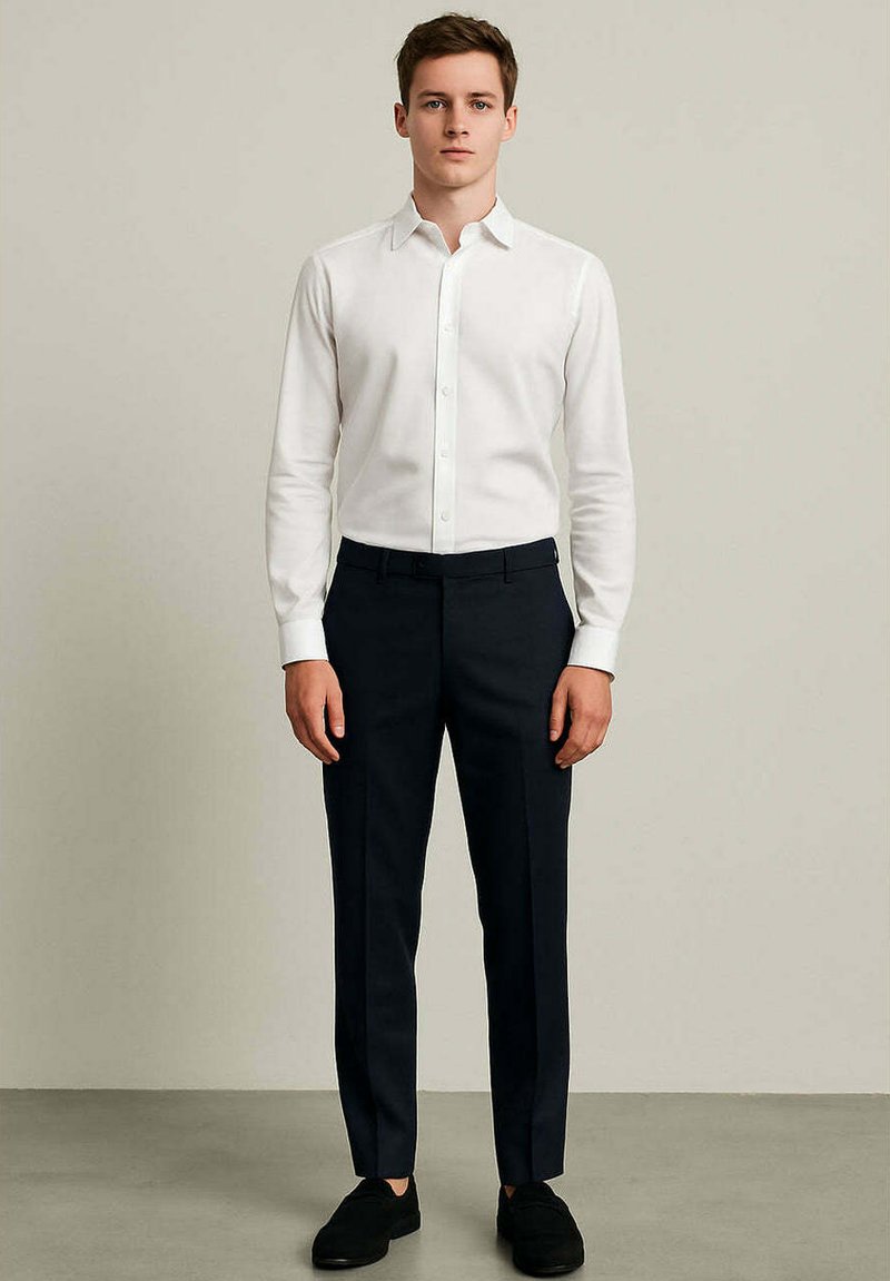 Camicia bianca a maniche lunghe con bottoni abbinata a pantaloni scuri e scarpe nere. La camicia è aderente; i pantaloni hanno un design su misura.