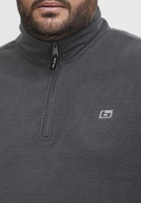 Grauer Fleece-Sweatshirt mit halblangem Reißverschluss, weichem Material und kleinem Logo auf der Brust. Der Reißverschluss verfügt über einen schwarzen Zieh-Tab.