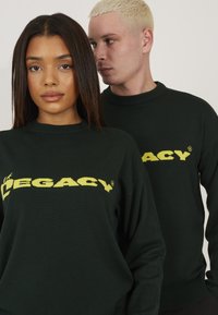 Dunkelgrüne Strick-Sweatshirts mit gelbem "LEGACY"-Logo über der Brust, gerippten Bündchen und Halsausschnitt, präsentiert von zwei Models.