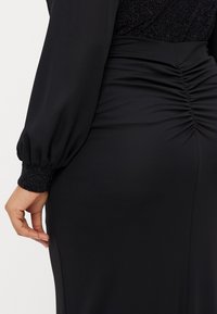 Svart kjole med lange puffærmer, rynket sidede, og teksturert svart mansjett. Laget av glatt stoff med skinnende detaljer.