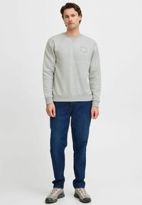 Sudadera gris claro con puños y bajo acanalados, con un detalle de logo redondo en el pecho. Combinada con jeans azul oscuro y zapatos grises.