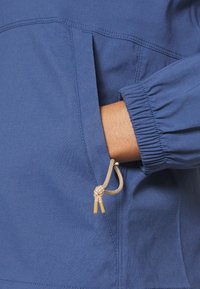 Main partiellement insérée dans la poche avant d'une veste bleue avec un poignet élastique et un cordon beige.