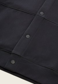 Felpa nera con una texture liscia, dettagli con bottoni a scatto e polsini a costine. Colore uniforme e design minimal.