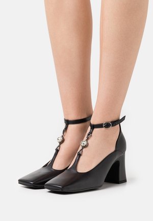 Chaussures à talons noires avec bout carré et bride à la cheville. Présentent un design en T et un accent métallique décoratif près de la cheville. Texture lisse.