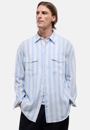 Homme aux cheveux mi-longs portant une chemise ample à rayures verticales bleu clair et blanc avec deux poches poitrine.