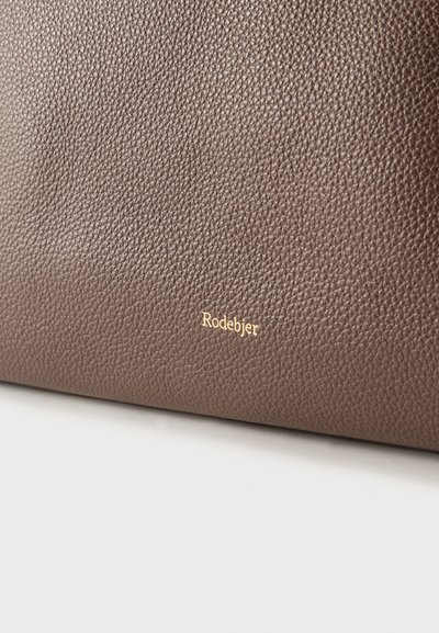 Gros plan sur une surface en cuir brun texturé avec un petit logo doré "Rodebjer" embossé près du bord inférieur.