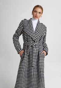 Czarnobiały wzór houndstooth, długi wełniany płaszcz z paskiem, szerokimi klapami i kieszeniami po obu stronach, z teksturowanym wykończeniem.