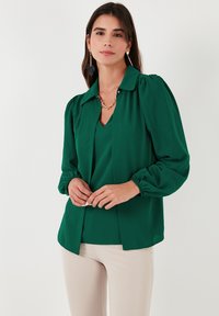 Blusa verde con cuello tipo camisa, mangas largas abullonadas y escote en V. Hecha de una tela ligera; combinada con pantalones beige.