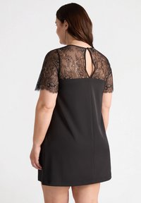 Schwarzes Kleid mit einer taillierten Taille und kurzen, durchsichtigen Spitzenärmeln. Detail mit Schlüssel-Ausschnitt auf der Rückseite, glatte Textur und oberschenkellange Länge.