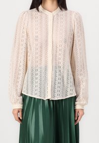 Lace blouse in cream with a high collar and button closure, featuring intricate floral patterns and long puffed sleeves.