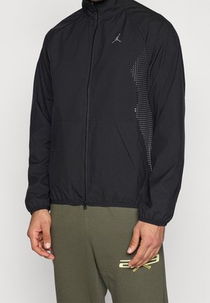 Homme portant une veste noire zippée avec des poignets élastiques et des accents latéraux à pois, associée à un pantalon vert avec un logo jaune.
