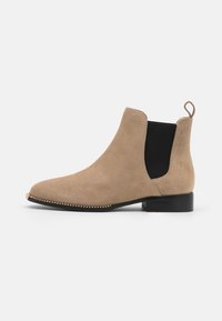 Botín de tobillo de ante beige con paneles laterales elásticos negros, punta redondeada, acento dorado a lo largo de la suela y una pequeña etiqueta en el talón.