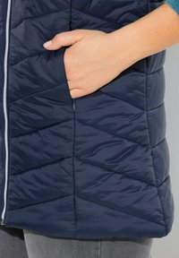 Marineblauem gepolsterten Weste mit Chevron-Nähten, ausgestattet mit einer Reißverschlusstasche und einer glatten, glänzenden Textur. Nahaufnahme einer Hand in der Tasche.