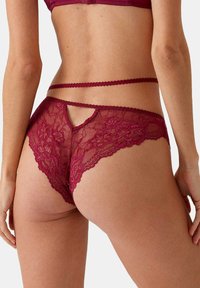 Bordeaux kanten string met bloemmotieven, doorschijnende gaasdetails en een sleutelgatopening aan de achterkant. Voorzien van dunne bandjes en geschulpte randen.