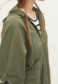 Olive green hooded jacket with zippered front, side pockets, and a textured finish. Worn over a striped shirt. Features silver hardware accents.